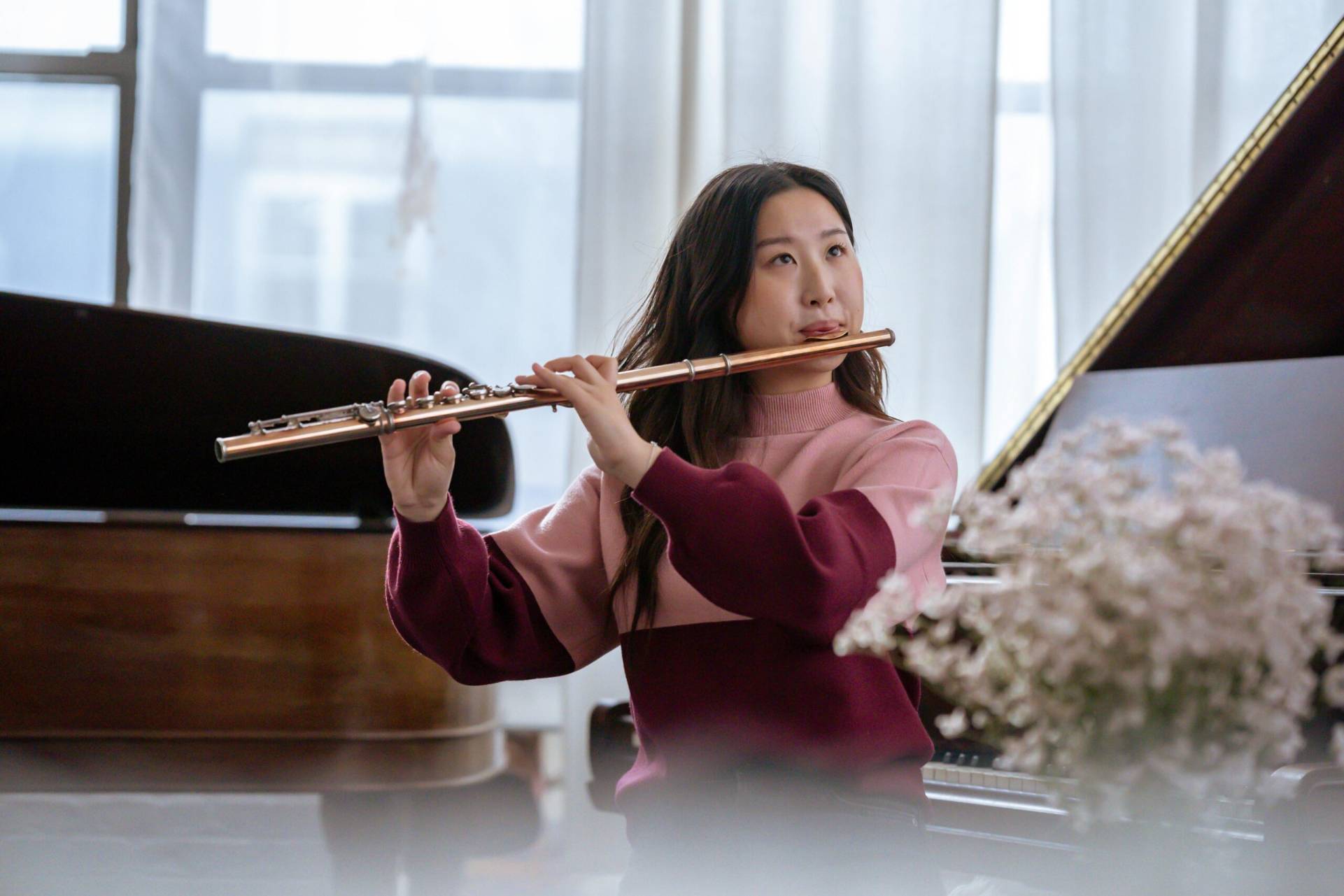 Asian woman plays a flute in a bright, serene room with a piano backdrop. Perfect for music hobbies.